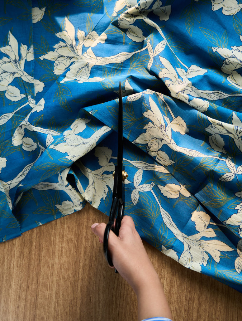 Person cutting blue floral fabric with scissors on a wooden surface